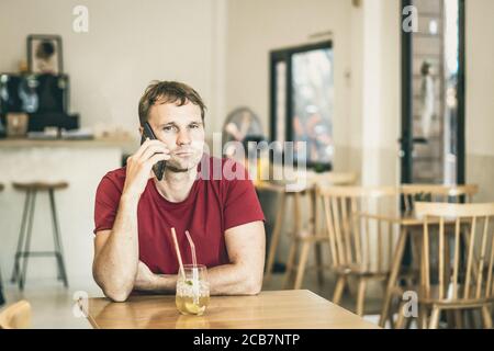 Ernst traurig junger Mann Beantwortung Smartphone in roten Hemd im Café trinken Eistee, warten jemand oder Coworking. Ausdruck echter Menschen, Leben Stockfoto