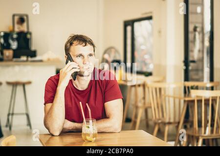 Ernst traurig junger Mann Beantwortung Smartphone in roten Hemd im Café trinken Eistee, warten jemand oder Coworking. Ausdruck echter Menschen, Leben Stockfoto