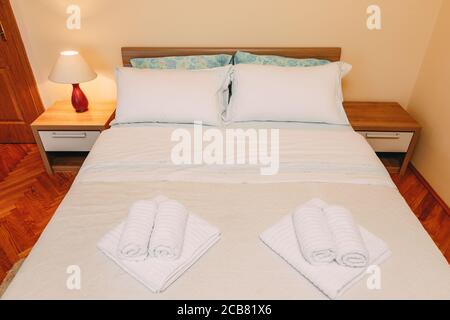 Großes Doppelbett mit Kissen und Frottee-Handtüchern Die Tagesdecke in einem geräumigen Zimmer mit einer Tischlampe Auf dem Nachttisch bei der Tür in einem Raum mit Stockfoto