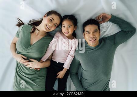 Asiatische Mutter und Vater liegen mit niedlichen Tochter auf dem Bett Blick auf Kamera und lächelnd. Glückliche Familie und Zweisamkeit Lifestyle-Konzept Stockfoto