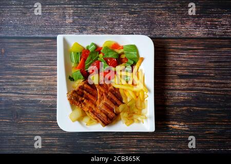Schweinesteak mit Pommes Frites und Gemüse (pak Choi, Zwiebeln, Tomaten, Paprika) auf einem weißen quadratischen Teller auf einem dunklen Holztisch, deutsche Hausmannskost. Stockfoto