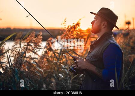 Kaukasischer bärtiger Mann in Hut Angeln im Schilf in der Nähe des Sees. Sonnenuntergang. Stockfoto