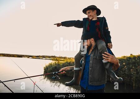 Vater und Sohn haben Spaß beim Angeln. Teen junge glücklich und lächeln, sitzen auf Vater Schultern und zeigt Hand auf Schwimmer Stockfoto