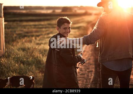 Vater und Sohn nahmen Angelruten und Angelausrüstung. Sie gehen auf Landstraße in der Nähe des Sees zum Angeln. Junge lächeln und schauen Sie sich die Kamera. Sonnenuntergang. Stockfoto