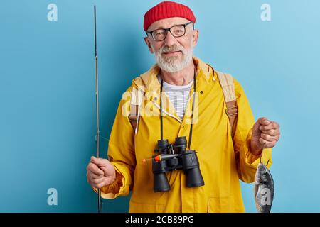 Freundliche gut aussehende bärtige Mann in stilvollen Freizeitkleidung hält Fisch und Stange poding an die Kamera, Lifestyle, Freizeit Stockfoto