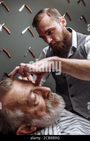 Nahaufnahme eines professionellen Friseursalon, der einen alten Mann in einem modernen Friseurladen rasiert. Europäischer älterer Mann, der seinen Bart vom Friseur rasiert Stockfoto