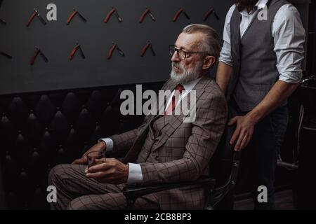Grauhaarige alte männliche Geschäftseigentümer in formeller Kleidung und optische Brille gekleidet, Anzug mit roter Krawatte, sitzen im Luxus Herren Schönheitssalon, Seitenansicht, Wel Stockfoto