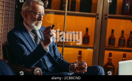 Stattliche alte männliche Rentner in Smart formalen Stil mit Laptop und Rauchen, während auf der Couch im Luxury Home Interior sitzen mit Schaltschrank mit ex gekleidet Stockfoto