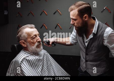 Nahaufnahme eines Friseur s Arbeiten für einen stattlichen respektables Alter-alter bärtiger Mann an der Friseur. Er tut Styling mit dem elektrischen Rasierer. Stockfoto