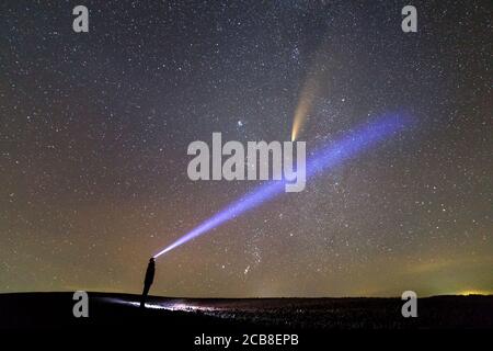 Silhouette eines Mannes mit Taschenlampe auf dem Kopf zeigt hellen Lichtstrahl auf Sternenhimmel mit C/2020 F3 (NEOWISE) Kometen mit Lichtschweif. Stockfoto