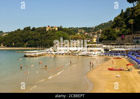 Erhöhter Blick auf einen Sandstrand mit Urlaubern in den Wellen des Ufers und einem bewaldeten Hügel im Hintergrund im Sommer, Lerici, La Spezia, Italien Stockfoto