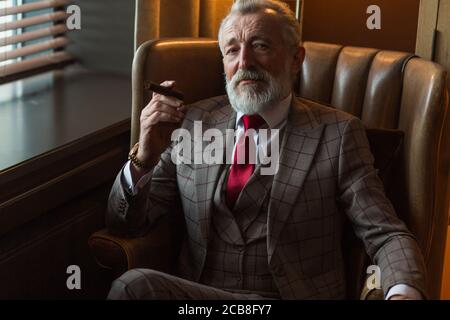 Ernsthafte alte alter Geschäftsmann tragen edlen maßgeschneiderten Anzug, roter Krawatte und teure Armbanduhr sitzen auf altmodische Sofa im Büro, Stockfoto