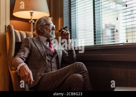 Ernsthafte alte alter Geschäftsmann tragen edlen maßgeschneiderten Anzug, roter Krawatte und teure Armbanduhr sitzen auf altmodische Sofa im Büro, Stockfoto