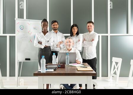 Porträt von lächelnden multirassischen jungen Geschäftsleuten in der Nähe von Chef, der Sitzen Sie am Schreibtisch im Büro und schauen Sie sich die Kamera an Stockfoto