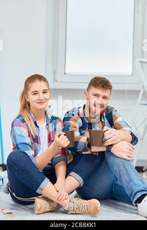 Glückliches Paar in karierten Hemden und Denim-Hosen sitzen auf dem Boden mit Tassen Kaffee, eine Pause von der Malerei ihrer neuen Wohnungen Stockfoto