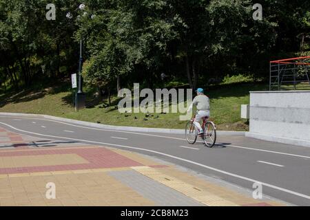 Chabarowsk, Russland - 20. August 2019. Ein älterer Mann in leichter Sportkleidung und blauer Mütze fährt in einem Stadtpark entlang eines markierten Radweges. Stockfoto