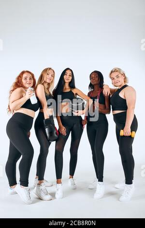 Fünf lächelnde multiethnische Frauen tragen sportliche Leggins und Themen halten Fitness-Ball, Boxhandschuhe und andere Dinge isoliert auf weißem Hintergrund Stockfoto