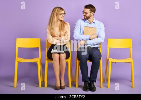 Lächelnder junger Mann mit Papieren und eine Mitarbeiterin mit gekreuzten Die Hände in der Brille sitzen und einander anschauen und Warten auf Bewerbungsgespräch Stockfoto