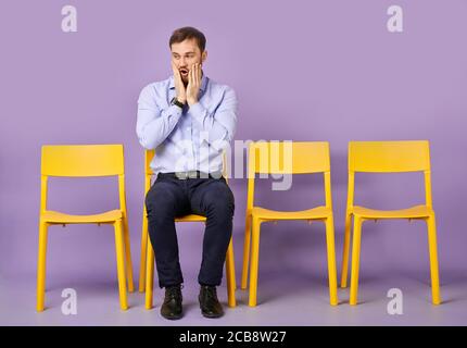 studioaufnahme des staunenden und staunenden jungen Mannes, der darauf sitzt Stuhl und Blick beiseite und warten auf Vorstellungsgespräch Stockfoto