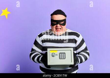 Glücklich fröhlich aufgeregt Einbrecher in Maske und gestreifte Kleidung hält Safe isoliert über blau. Close up Foto. Stockfoto