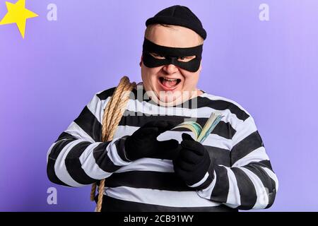 Fröhlicher Räuber in gestickter Kleidung ist glücklich, wie er viel Geld gestohlen hat. Nahaufnahme Foto. Isoliert blauen Hintergrund. Stockfoto