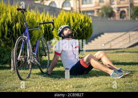 Fröhlicher junger bärtiger Radfahrer sitzt auf Gras Stockfoto