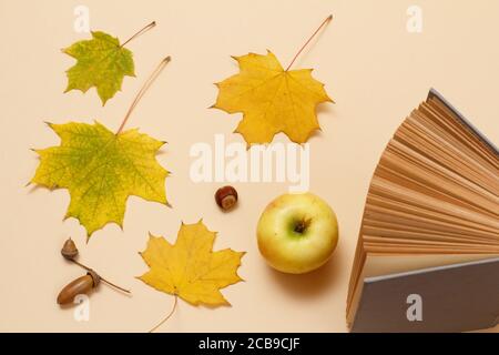 Reifer Apfel, ein Buch, trockene gelbe Ahornblätter und Eicheln auf beigem Hintergrund. Draufsicht. Stockfoto