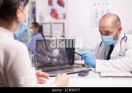 Arzt zeigt auf Coronovirus-Darstellung auf einem Laptop-Display und informiert Patienten über die COVID-19-Krankheit Stockfoto
