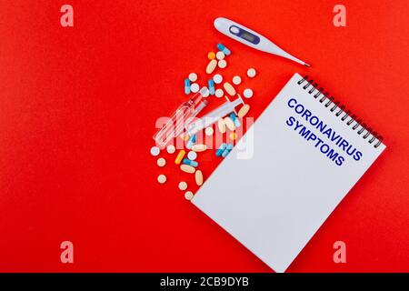 Notizblock, eine Vielzahl von Medikamenten, Thermometer, Spritze und Ampullen mit Medizin auf rotem Hintergrund. In blau auf einem Notizblock CORONAVIRUS-Symbtome geschrieben. Stockfoto