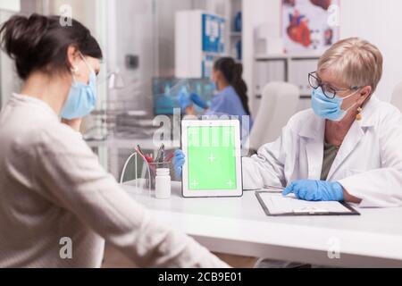 Arzt mit Gesichtsmaske gegen Coronavirus hält Tablet-Computer mit grünem Bildschirm während der Konsultation mit kranken Patienten. Stockfoto