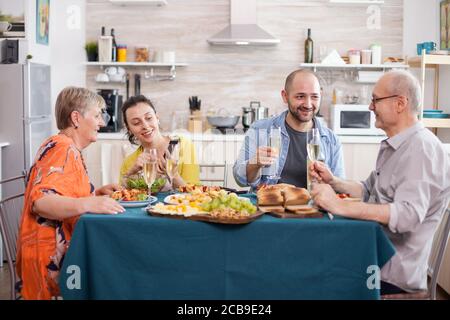 Vater und Sohn halten Weingläser während des Familienbrunchs. Tochter und ältere Mutter betrachten Smartphone. Leckere gewürzte Kartoffeln. Stockfoto
