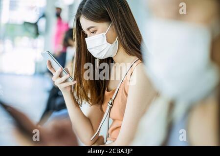 Junge asiatische Frau Passagier trägt chirurgische Maske und mit mobilen Telefon für soziales Netzwerk beim Warten Bus am Bahnhof mit Gekrönte Menschen, wenn trave Stockfoto