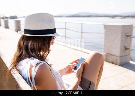 Kaukasische junge Frau mit weißem Hut auf einer Bank sitzen Eines Gehwegs am Meer mit ihrem Smartphone - Single Frau im Freien mit neuen sozialen verbunden Stockfoto
