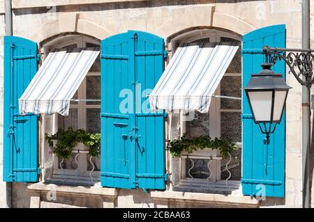 Zwei Fenster mit blauen hölzernen Fensterläden und gestreiften Markisen, Topfblumen auf dem Boden. Eine Straßenlaterne auf der rechten Seite. Stockfoto