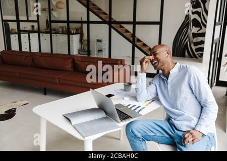 Foto eines lachenden afroamerikanischen Mannes, der während der Verwendung des kabellosen Ohrhörers den Hörer benutzte Arbeiten mit Laptop am Tisch im Wohnzimmer Stockfoto