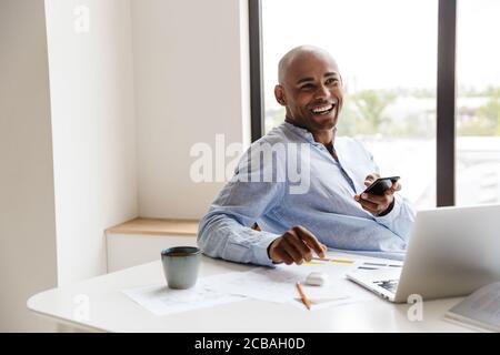 Foto von lachenden afroamerikanischen Mann mit Handy während der Arbeit Mit Laptop am Tisch im Wohnzimmer Stockfoto