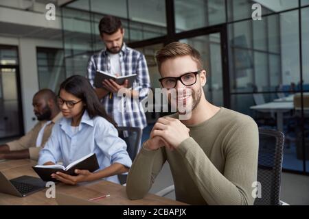 Porträt eines jungen kaukasischen männlichen Büroangestellten mit Brillen Am Schreibtisch sitzend und beim Arbeiten lächelnd auf die Kamera Zusammen mit Stockfoto