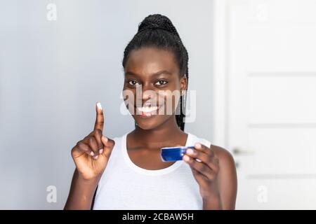 Close up Kopf Schuss lächelnd afroamerikanische junge Frau Anwendung feuchtigkeitsspendende Gesichtscreme oder Maske auf Wange nach der Dusche, attraktive Mädchen tragen weiß Stockfoto