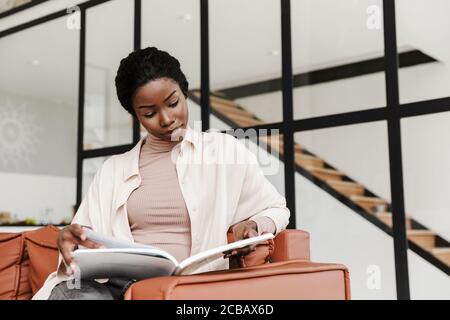 Attraktive junge afrikanische Frau sitzt auf einer Couch zu Hause, moderne Innenausstattung, ein Buch zu lesen Stockfoto