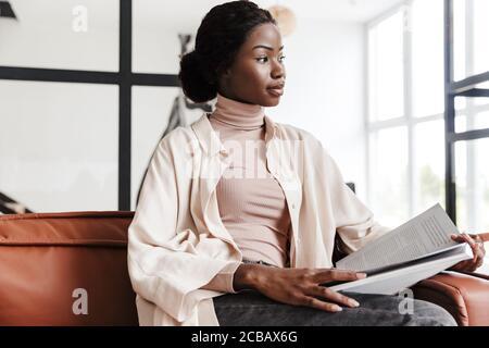 Foto einer ernsthaften jungen afrikanischen Frau, die drinnen auf dem Sofa sitzt Zu Hause beim Lesen von Zeitschriften oder Büchern Stockfoto