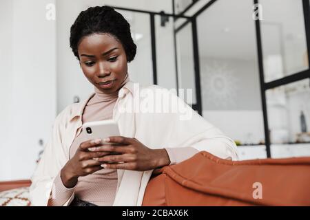 Bild einer ernsthaften jungen afrikanischen Frau, die drinnen auf dem Sofa sitzt Zu Hause, während Sie das Mobiltelefon verwenden Stockfoto