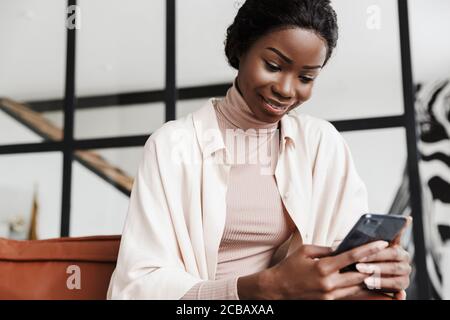 Attraktive junge lächelnde afrikanische Frau sitzt auf einer Couch zu Hause, moderne Einrichtung, mit Handy Stockfoto