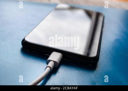 Weißes Telefonladegerät, das an einem blauen Schreibtisch zu Hause an ein Smartphone angeschlossen ist. Intelligente Technik und Ladekonzept Stockfoto