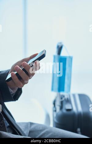 Geschäftsmann, in grauem Anzug, sitzt in der Wartehalle eines Flughafens oder eines Bahnhofs, mit seinem Smartphone, und eine chirurgische Maske hängen von der te Stockfoto