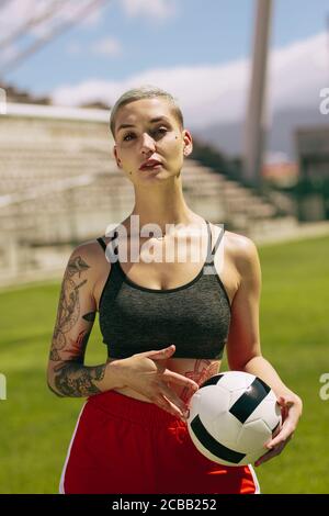 Porträt einer Fußballspielerin, die einen Ball auf dem Fußballfeld hält. Frau hält einen Ball und schaut mit der Kamera auf das Fußballfeld Stockfoto