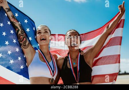 Weibliche Athleten feiern den Sieg mit amerikanischer Flagge im Stadion. Fröhliche Läuferinnen schreien vor Freude und halten die US-Flagge nach winni Stockfoto