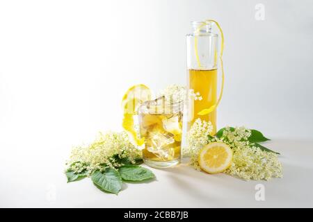 Holunderblütensirup in einer Flasche und hausgemachte Limonade mit Eiswürfeln in einem Trinkglas, Zitronenschale und Scheiben und einige frische Blüten auf einem Licht Stockfoto