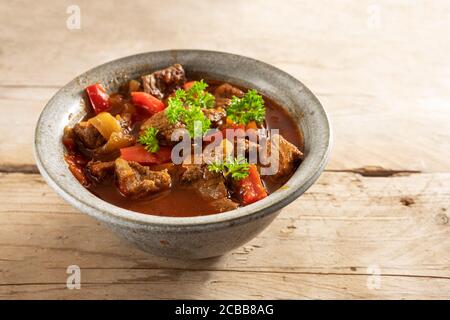 Frisch gegartes Gulasch oder Eintopf aus Rindfleisch, Zwiebeln und rotem Paprika mit Petersilie garnieren in einer Schüssel auf einem rustikalen Holztisch, Kopierraum, ausgewählter Fokus Stockfoto