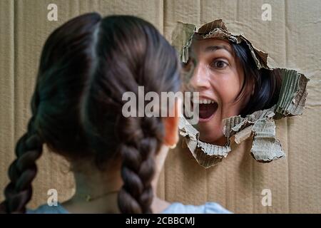 Frau mit offenem Mund, die ein Mädchen durch ein großes Loch in einem Karton anschaut. Stockfoto