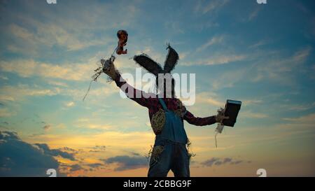Der Mann im Bild des Zauberers im Hut mit Kaninchenohren führt ein Voodoo-Ritual durch und hält Puppe mit aufgespießenem Draht gegen den Sonnenuntergang am Himmel. Stockfoto